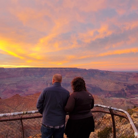 Two people at a Grand Canyon viewpoint during a colorful sunset.