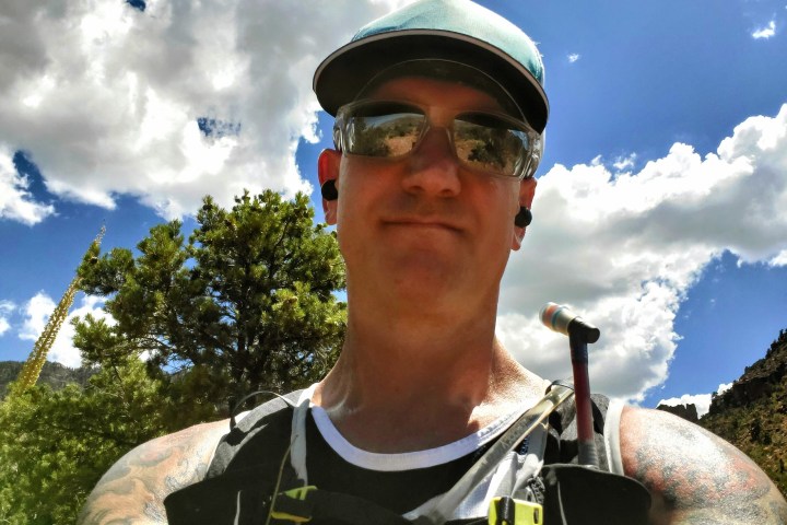 Person hiking outdoors, wearing sunglasses and a cap, with clouds and trees in the background.