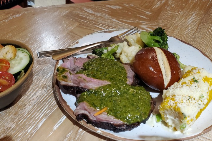 Plate with meat, pesto, veggies, corn on the cob, pretzel roll; salad with tomatoes and croutons on the side.