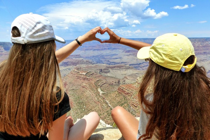 Two people sit on a cliff edge, making a heart shape with hands, overlooking the Grand Canyon.