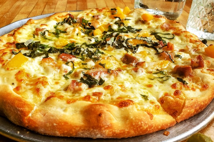 Close-up of a pizza with cheese, yellow peppers, and basil on a metal tray, with drinks in the background.