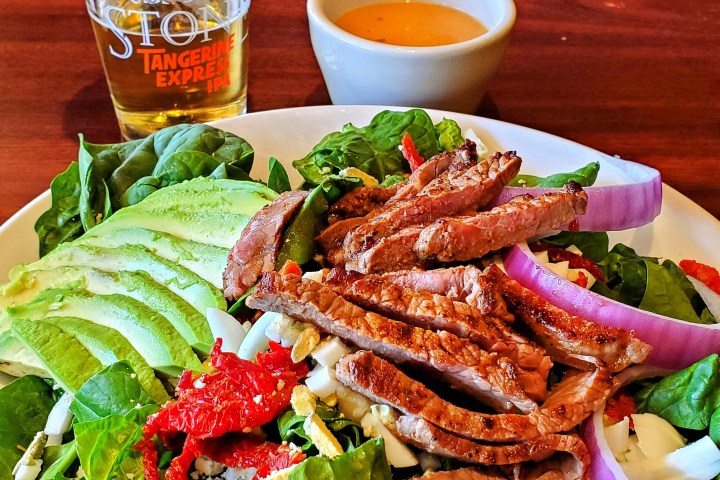 Salad with grilled meat, avocado, and onions, a beer, and a dipping sauce on a wooden table.