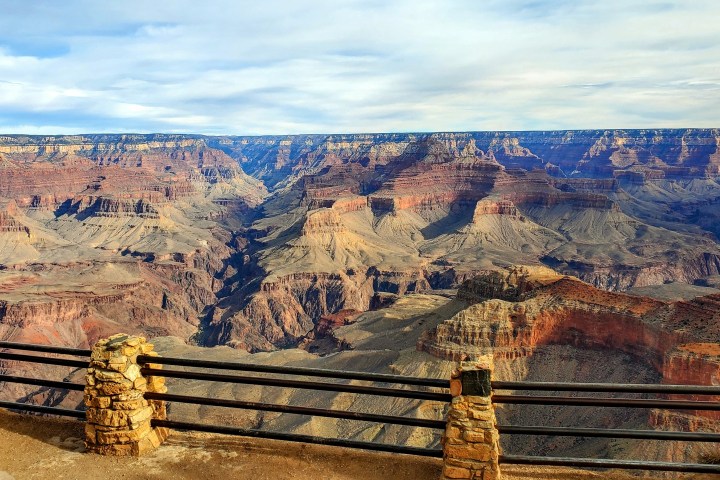 View of the Grand Canyon with stone wall and railing in the foreground under a partly cloudy sky.